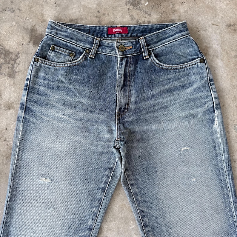 May include: Faded blue denim jeans with a high waist and classic five-pocket design. The jeans have a button and zipper closure, with a small red tag that reads "SOMETHING" above the back pocket. The denim has a worn, distressed look.