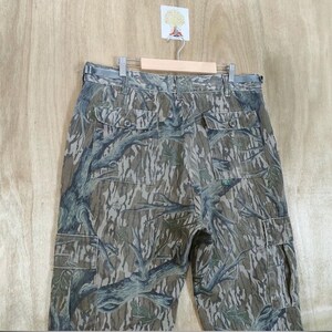 May include: Camouflage cargo trousers with multiple pockets, hanging on a wooden hanger. The trousers feature a pattern of brown, green, and grey tree branches and leaves. The trousers have a button and zip closure and a belt loop.
