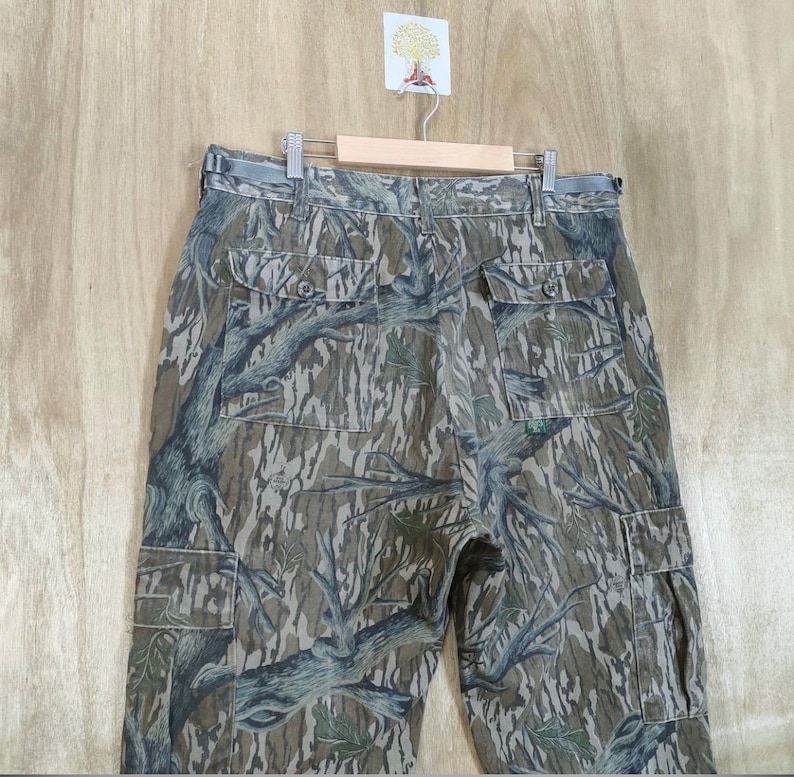 May include: Camouflage cargo trousers with multiple pockets, hanging on a wooden hanger. The trousers feature a pattern of brown, green, and grey tree branches and leaves. The trousers have a button and zip closure and a belt loop.