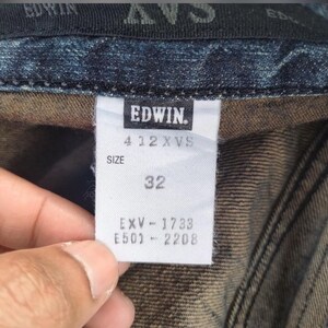 Może przedstawiać: Zbliżenie białej metki z dżins&oacute;w Edwin. Na metce widnieje nazwa marki, numer stylu 412 XVS oraz rozmiar 32. Dodatkowe kody EXV-1733 i E501-2208 są r&oacute;wnież wydrukowane na metce. Metka jest przymocowana do ciemnoniebieskiego denimu.