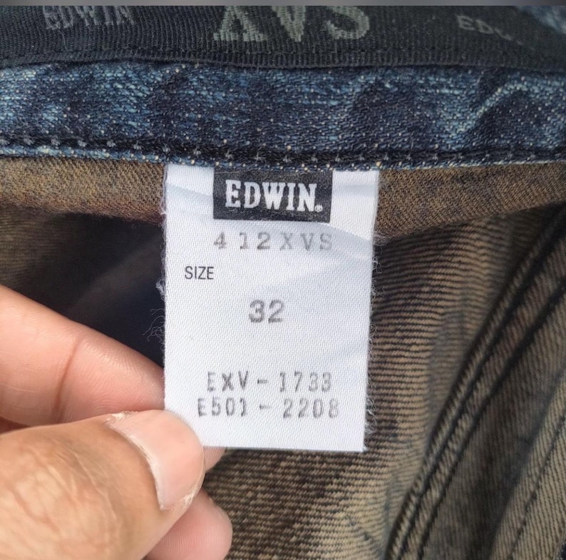 Może przedstawiać: Zbliżenie białej metki z dżins&oacute;w Edwin. Na metce widnieje nazwa marki, numer stylu 412 XVS oraz rozmiar 32. Dodatkowe kody EXV-1733 i E501-2208 są r&oacute;wnież wydrukowane na metce. Metka jest przymocowana do ciemnoniebieskiego denimu.