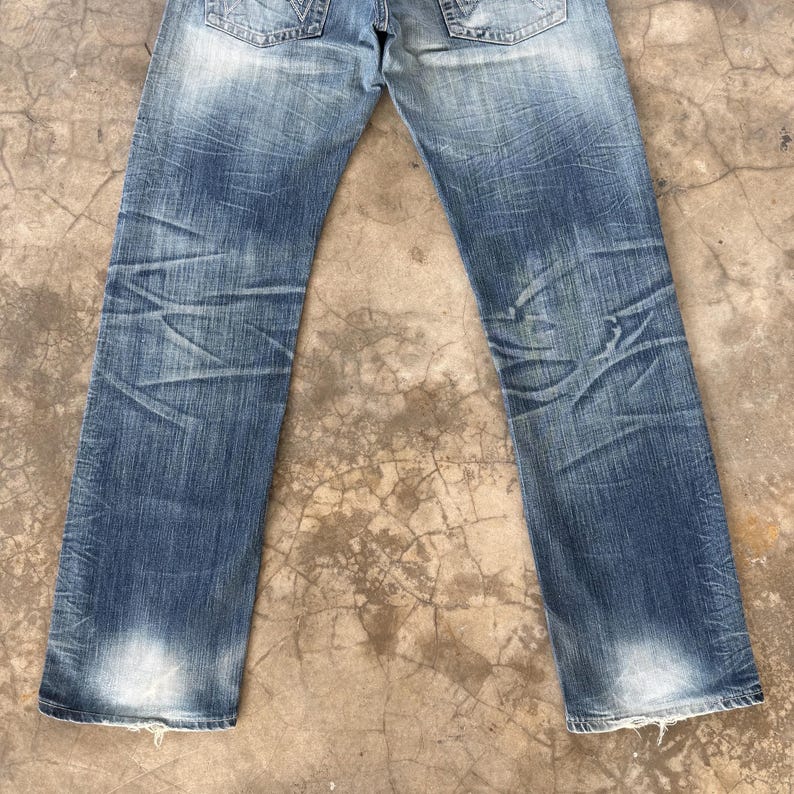 May include: Faded blue denim jeans with distressed detailing. The jeans have a classic five-pocket design, a button and zipper closure, and a worn appearance. The denim shows fading and wear patterns, with a light wash at the bottom of the legs.