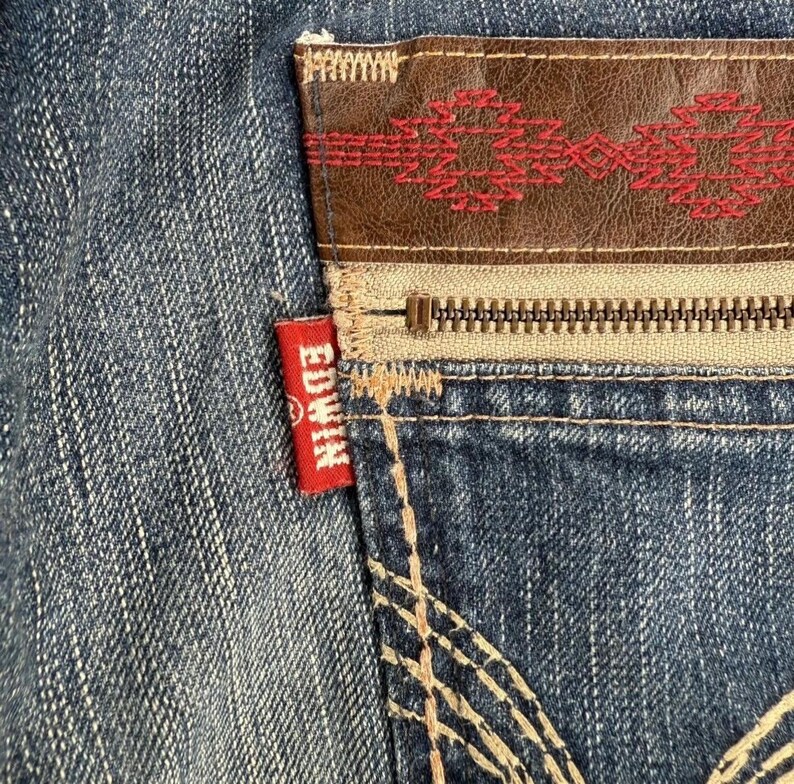 May include: Close-up of a denim jeans pocket with a brown leather patch featuring red embroidery. A silver zipper and the red "EDWIN" tag are visible. The jeans have a faded blue wash.
