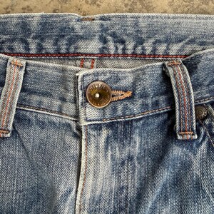 May include: Close-up of blue denim jeans. The jeans have a button closure and belt loops. The stitching is orange and the denim has a faded appearance. The jeans are a classic style.