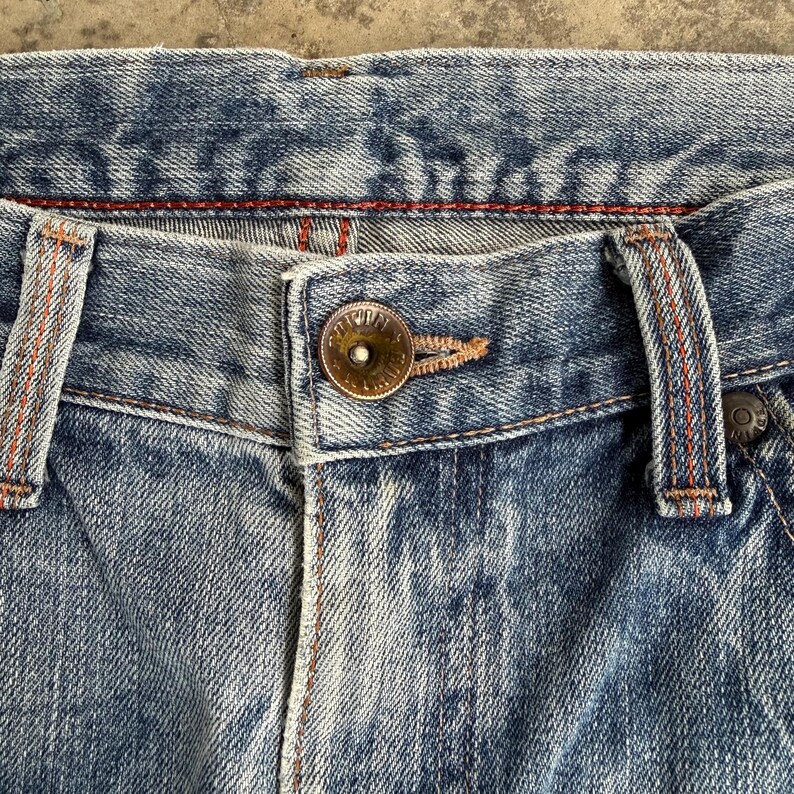 May include: Close-up of blue denim jeans. The jeans have a button closure and belt loops. The stitching is orange and the denim has a faded appearance. The jeans are a classic style.