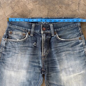 May include: Close-up of blue denim jeans with a button closure and a small coin pocket. The waist measures approximately 16 inches across. The jeans show signs of wear and fading, with a distressed look.