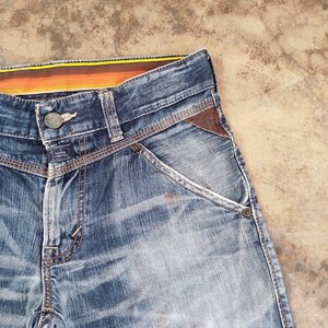 以下が含まれることがあります： ボタン留めと茶色の革のアクセントが付いた、青いデニムジーンズのクローズアップ。ジーンズは色あせたウォッシュ加工で、カラフルなストライプのウエストバンドが付いています。ポケットの縁は白いステッチでトリミングされています。