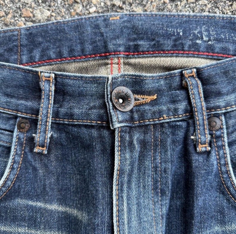 Pu&ograve; includere: Primo piano di jeans blu. I jeans hanno una chiusura con bottoni, passanti per cintura e tasche. Le cuciture sono di colore arancione a contrasto e i jeans sembrano usurati e sbiaditi.