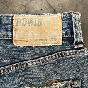 Puede incluir: Primer plano de unos vaqueros de mezclilla azul con una etiqueta de cuero color beige. La etiqueta rectangular tiene la marca "EDWIN" impresa. Los vaqueros tienen costuras visibles y un aspecto desgastado.