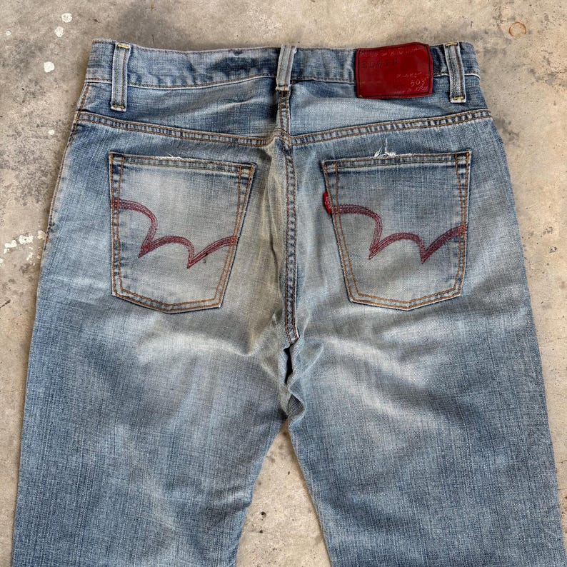 May include: Light blue denim jeans with red stitching on the back pockets. The jeans have a faded appearance and a red leather patch on the back waistband. The pockets have a unique wave-like stitch design.