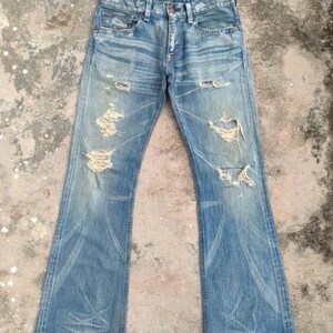 May include: Faded blue denim jeans with distressed details, including rips and worn patches. The jeans have a flared leg design and classic five-pocket styling. The denim shows signs of wear and fading.