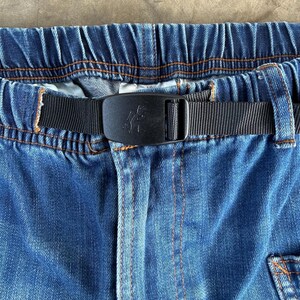 May include: Close-up of blue denim pants with an elastic waistband and a black nylon belt. The belt features a black buckle with a decorative design. Orange stitching accents the denim.