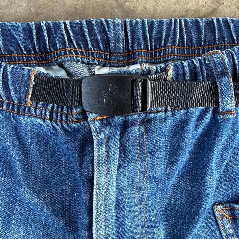 May include: Close-up of blue denim pants with an elastic waistband and a black nylon belt. The belt features a black buckle with a decorative design. Orange stitching accents the denim.