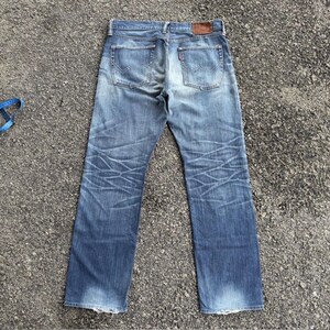 May include: Faded blue denim jeans with a classic five-pocket design. The jeans show wear and tear with faded areas and crease marks. A brown leather patch is visible on the back waistband, and a red tag is on the back pocket.