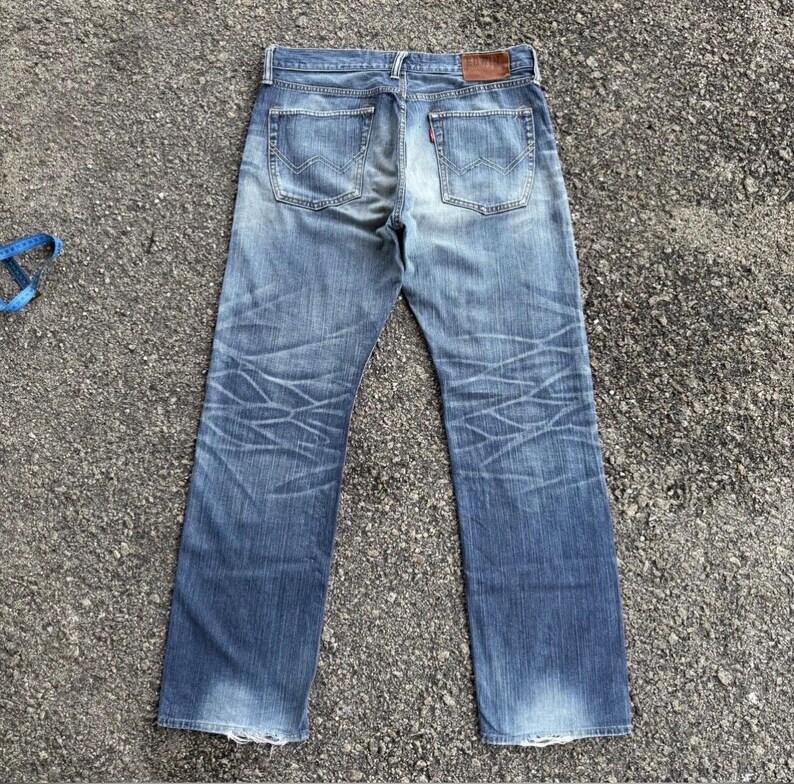 May include: Faded blue denim jeans with a classic five-pocket design. The jeans show wear and tear with faded areas and crease marks. A brown leather patch is visible on the back waistband, and a red tag is on the back pocket.