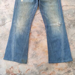May include: Faded blue denim jeans with a distressed appearance. The jeans feature a flared leg, a worn look, and a ripped section on the right knee. Orange stitching highlights the seams.