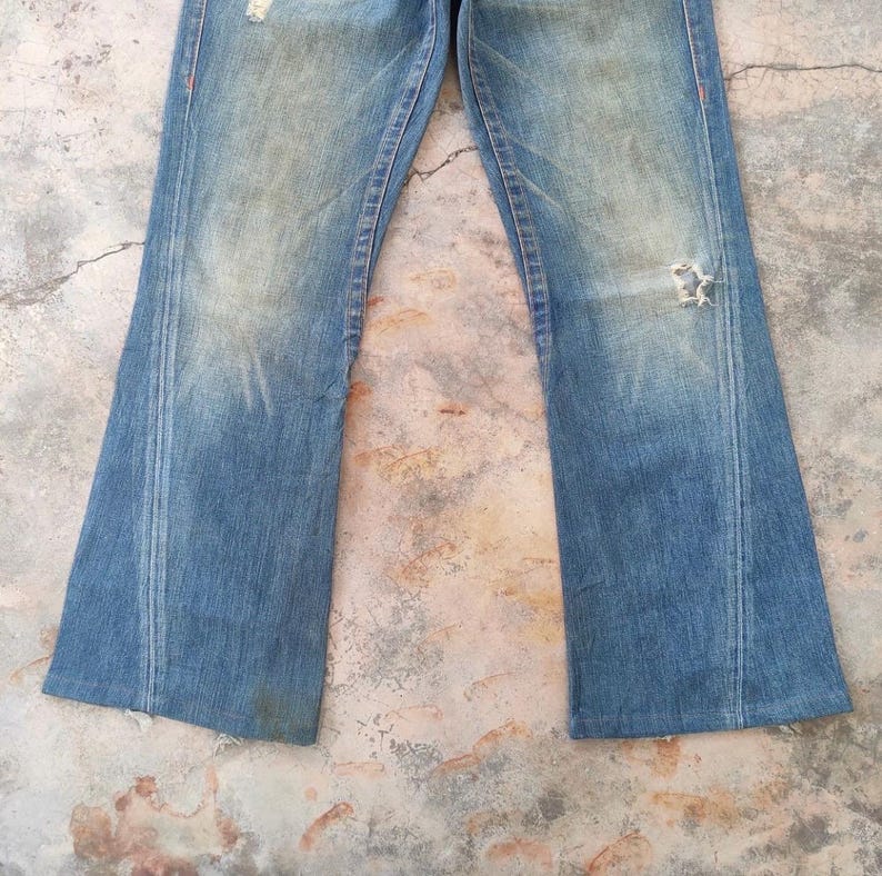 May include: Faded blue denim jeans with a distressed appearance. The jeans feature a flared leg, a worn look, and a ripped section on the right knee. Orange stitching highlights the seams.