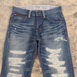 May include: Faded blue denim jeans with distressed details, including ripped sections and frayed edges. The jeans feature a classic five-pocket design, a button closure, and a decorative patterned waistband. The denim has a worn, vintage look.