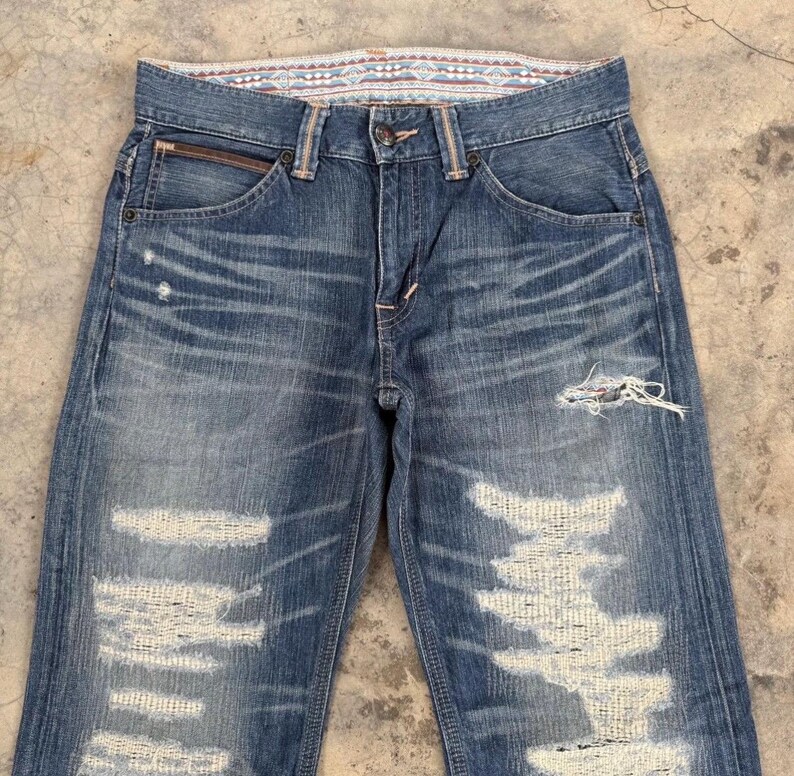 May include: Faded blue denim jeans with distressed details, including ripped sections and frayed edges. The jeans feature a classic five-pocket design, a button closure, and a decorative patterned waistband. The denim has a worn, vintage look.