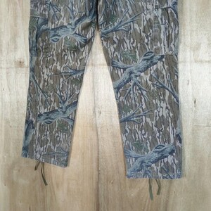 May include: Camouflage trousers with a tree branch pattern in shades of green, brown, and grey. The trousers have pockets and drawstrings at the bottom of the legs. The fabric has a textured appearance.