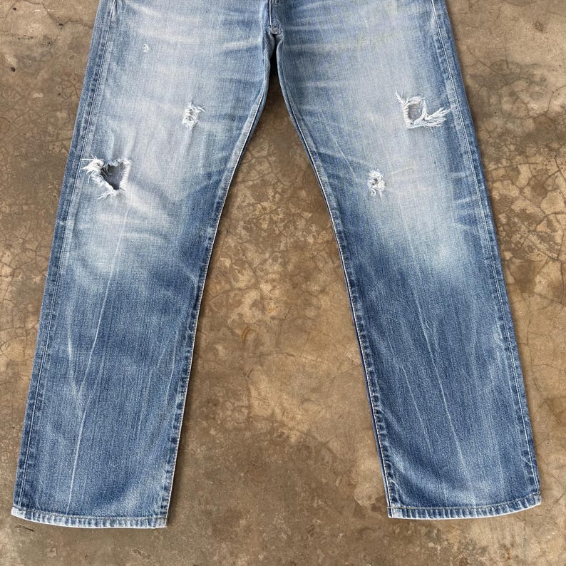 Pu&ograve; includere: Jeans di denim blu sbiadito con dettagli strappati, tra cui strappi e toppe usurate. I jeans hanno un classico design a cinque tasche e un taglio dritto. Il denim mostra segni di usura e sbiadimento.