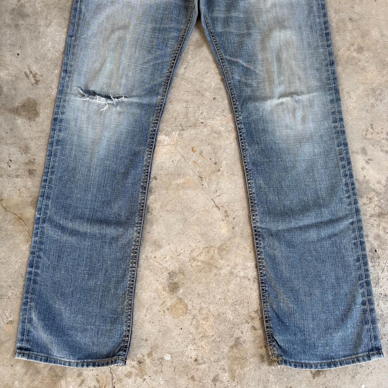 May include: Faded blue denim jeans with a distressed look. The jeans have a ripped knee and a classic five-pocket design. The denim has a worn appearance, with lighter areas and subtle fading throughout the fabric.