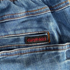 May include: Close-up of blue denim fabric with visible stitching in a contrasting orange-brown color. A black label with the red text "GRAMICCI" is sewn onto the denim. The fabric appears to be from a pair of jeans or similar garment.