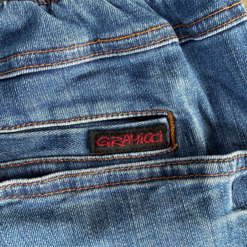 May include: Close-up of blue denim fabric with visible stitching in a contrasting orange-brown color. A black label with the red text "GRAMICCI" is sewn onto the denim. The fabric appears to be from a pair of jeans or similar garment.