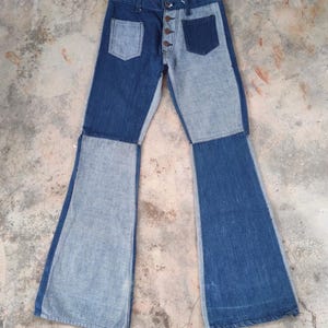 May include: Two-tone denim jeans with a button fly and two front pockets. The jeans feature a patchwork design with light and dark blue denim panels. The legs flare out into a bell-bottom style.