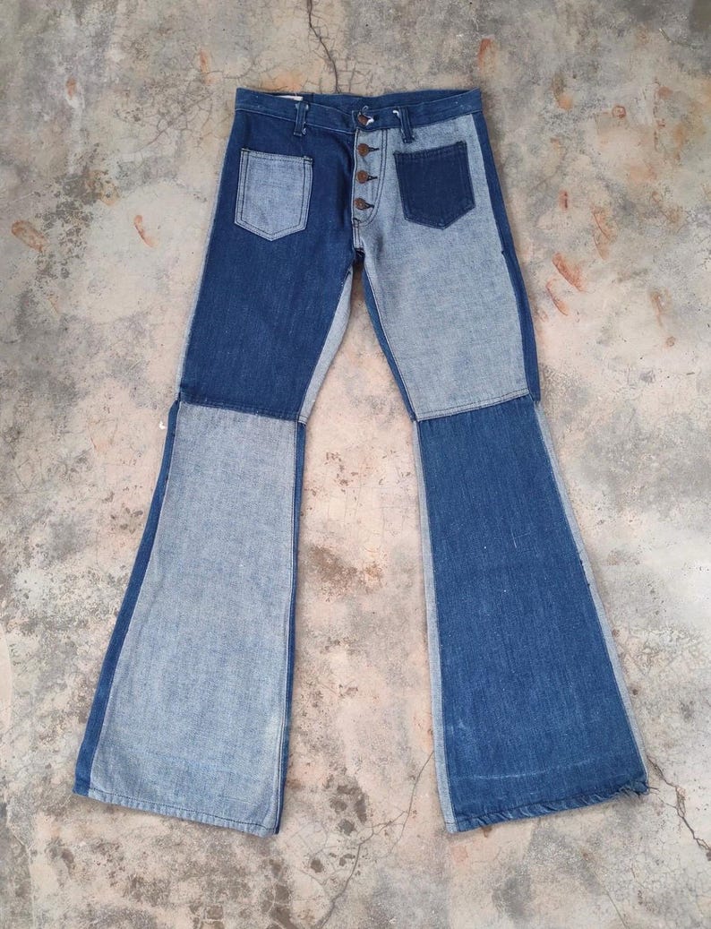 May include: Two-tone denim jeans with a button fly and two front pockets. The jeans feature a patchwork design with light and dark blue denim panels. The legs flare out into a bell-bottom style.