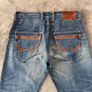 Pu&ograve; includere: Jeans in denim blu sbiadito con tasche con cerniera e cuciture arancioni. Le tasche posteriori presentano un esclusivo design a onda. Il nome del marchio "EDWIN" &egrave; visibile su un'etichetta in pelle marrone.
