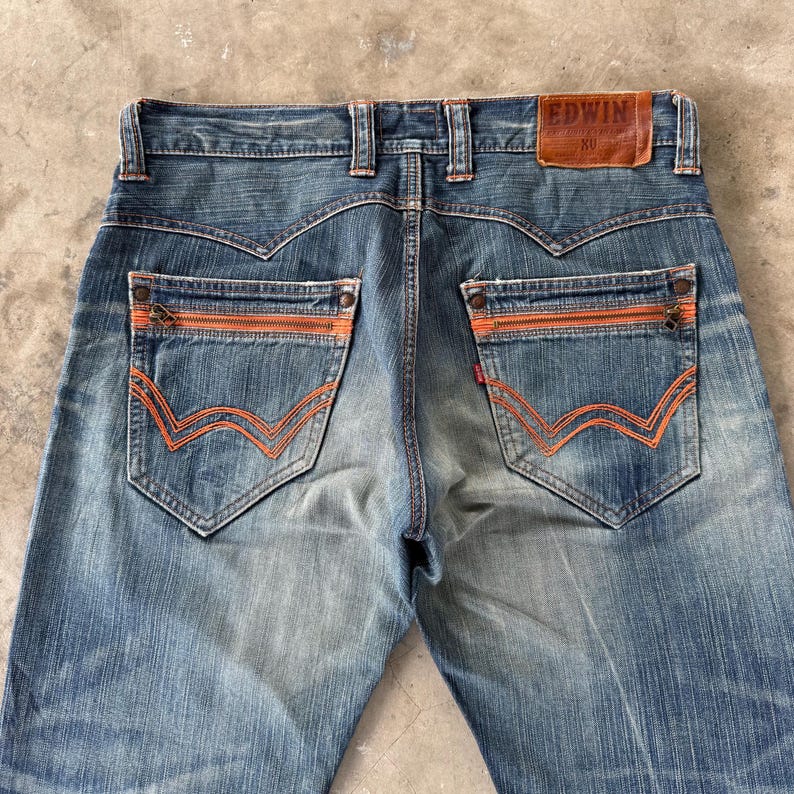 Pu&ograve; includere: Jeans in denim blu sbiadito con tasche con cerniera e cuciture arancioni. Le tasche posteriori presentano un esclusivo design a onda. Il nome del marchio "EDWIN" &egrave; visibile su un'etichetta in pelle marrone.