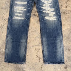 May include: Faded blue denim jeans with distressed detailing. The jeans feature ripped sections with white thread accents, creating a worn and vintage aesthetic. The straight-leg cut and subtle fading add to the casual style.