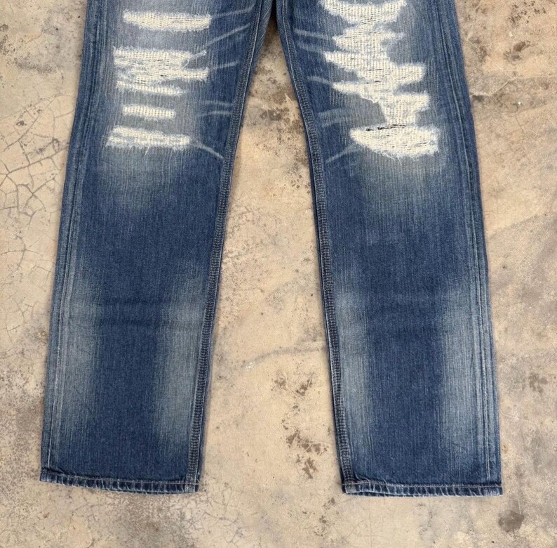 May include: Faded blue denim jeans with distressed detailing. The jeans feature ripped sections with white thread accents, creating a worn and vintage aesthetic. The straight-leg cut and subtle fading add to the casual style.