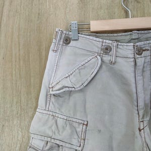 May include: Close-up of light beige cargo pants with multiple pockets and button details. The pants are hanging on a wooden hanger. The stitching is a darker beige color.