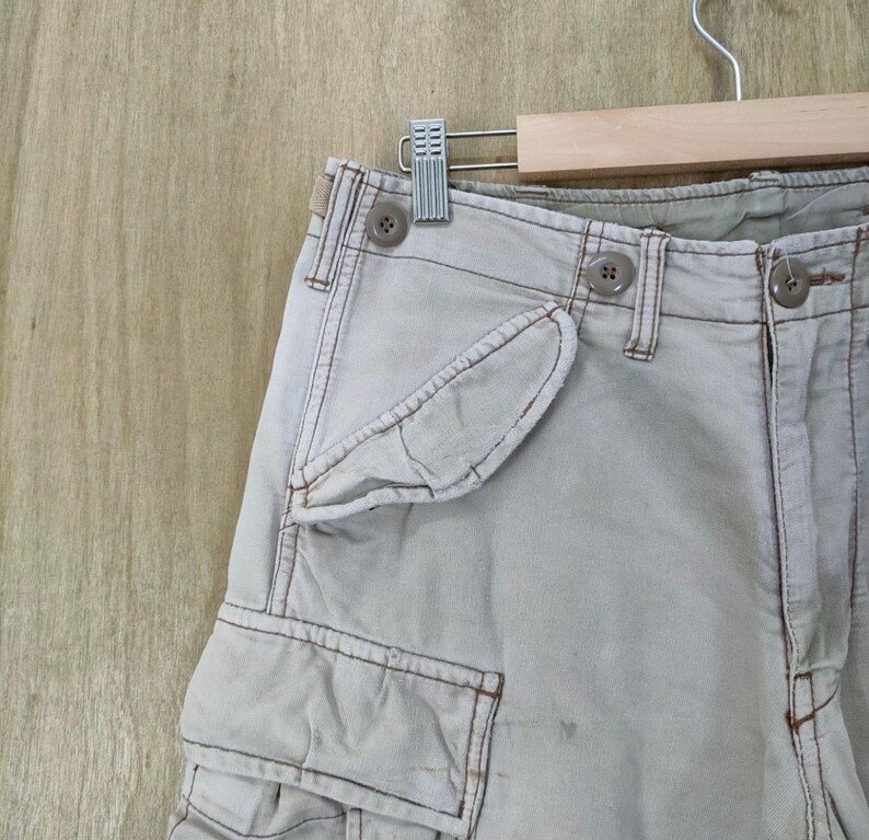 May include: Close-up of light beige cargo pants with multiple pockets and button details. The pants are hanging on a wooden hanger. The stitching is a darker beige color.