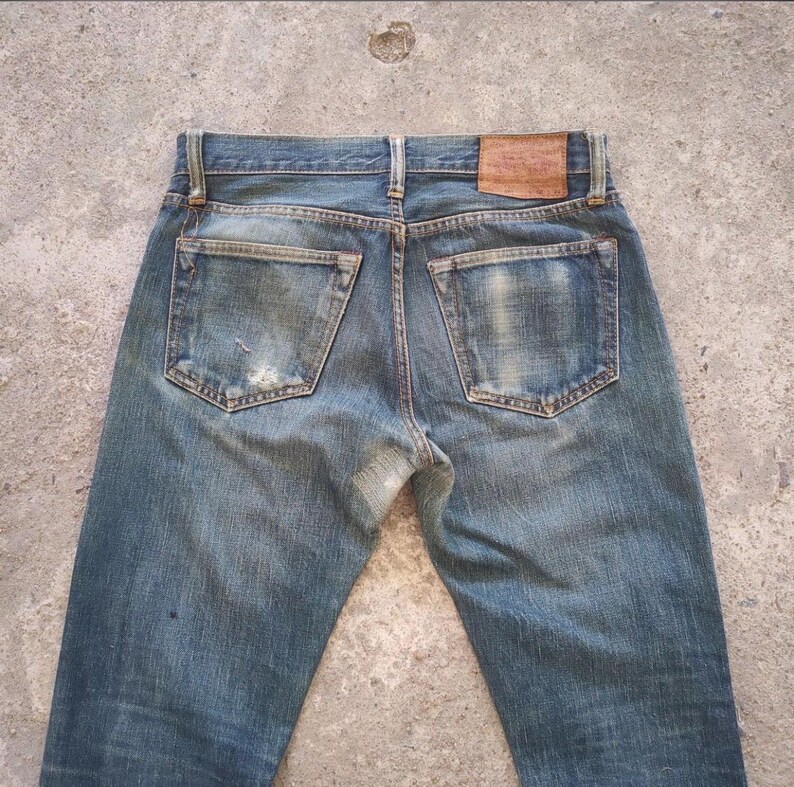 May include: Faded blue denim jeans with a worn appearance. The jeans feature two back pockets, a leather patch, and visible wear and tear. The jeans are a classic style.
