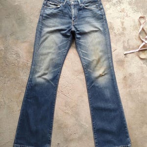 May include: Faded blue denim jeans with a flared leg. The jeans show signs of wear and tear, including a distressed area on the right knee. The jeans have classic five-pocket styling and a button and zip closure.
