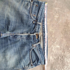 May include: Light blue denim jeans with a classic five-pocket design. The jeans are measured with a tape measure, showing a length of approximately 17 inches. The denim has a faded, worn look.