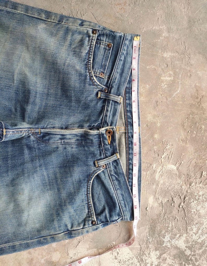 May include: Light blue denim jeans with a classic five-pocket design. The jeans are measured with a tape measure, showing a length of approximately 17 inches. The denim has a faded, worn look.