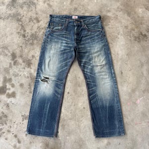 Pu&ograve; includere: Jeans in denim blu sbiadito con dettagli strappati, tra cui un ginocchio strappato. I jeans hanno un classico design a cinque tasche, una chiusura con bottoni e cuciture rosse. Il marchio &egrave; visibile.