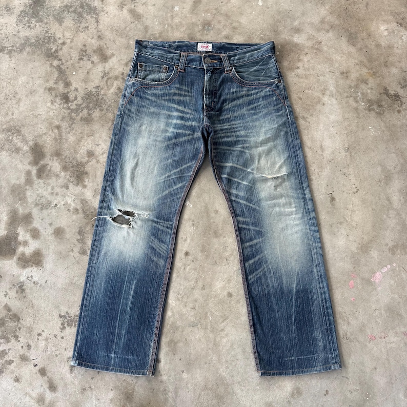Pu&ograve; includere: Jeans in denim blu sbiadito con dettagli strappati, tra cui un ginocchio strappato. I jeans hanno un classico design a cinque tasche, una chiusura con bottoni e cuciture rosse. Il marchio &egrave; visibile.