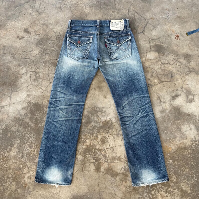 May include: Faded blue denim jeans with decorative back pockets and a red accent. The jeans have a worn appearance with distressed details and a light wash on the legs. A small tag is visible on the back.