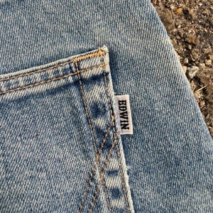 May include: Close-up of a light blue denim pocket with visible stitching and a white tag that reads "EDWIN". The denim has a textured appearance.