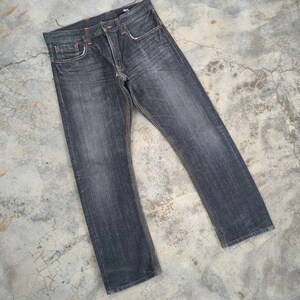Pu&ograve; includere: Jeans in denim grigio scuro con un classico design a cinque tasche. I jeans hanno un taglio dritto e presentano cuciture arancioni. Il tessuto mostra segni di usura, conferendo loro un aspetto vintage. I jeans sono stesi su una superficie di cemento.