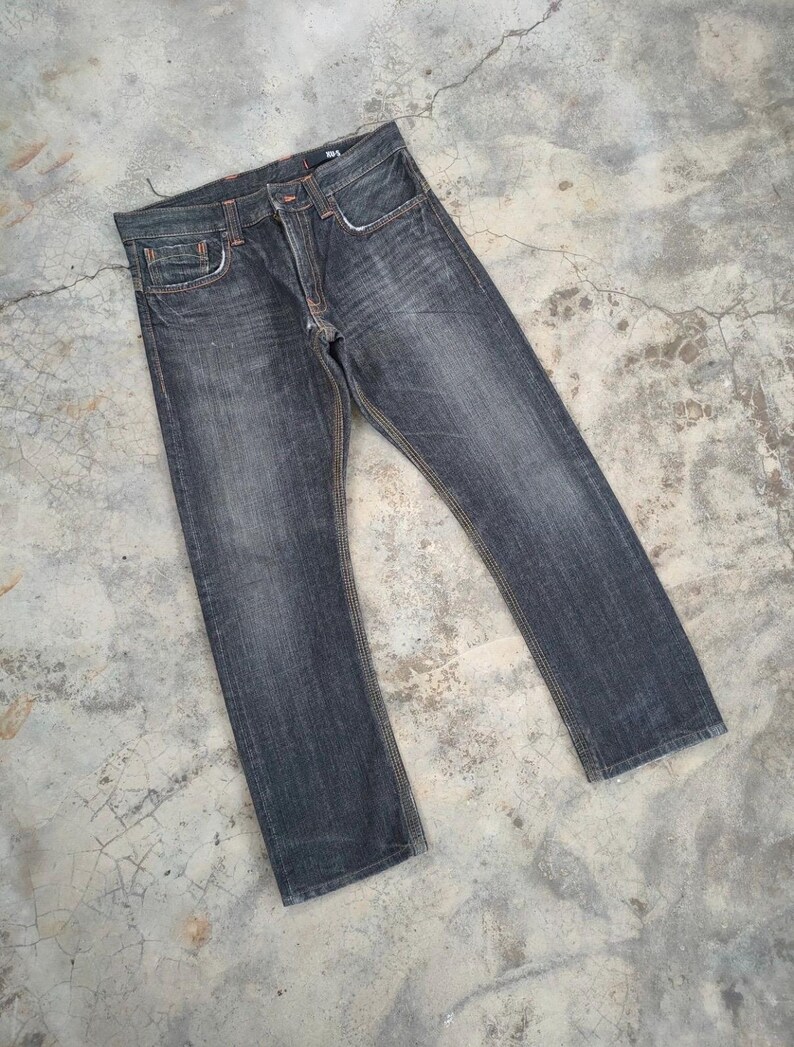 Pu&ograve; includere: Jeans in denim grigio scuro con un classico design a cinque tasche. I jeans hanno un taglio dritto e presentano cuciture arancioni. Il tessuto mostra segni di usura, conferendo loro un aspetto vintage. I jeans sono stesi su una superficie di cemento.