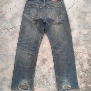 May include: Faded blue denim jeans with distressed details, including rips at the knees. The jeans feature classic five-pocket styling, a leather patch at the back, and a straight leg fit. The jeans are pre-worn and have a vintage aesthetic.