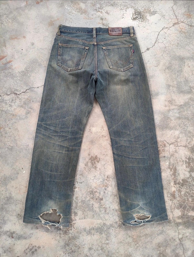 May include: Faded blue denim jeans with distressed details, including rips at the knees. The jeans feature classic five-pocket styling, a leather patch at the back, and a straight leg fit. The jeans are pre-worn and have a vintage aesthetic.