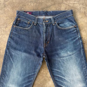 May include: Dark blue denim jeans with a classic five-pocket design. The jeans have a button and zipper closure, with copper-colored rivets and stitching. The denim has a faded appearance, with subtle wear marks.