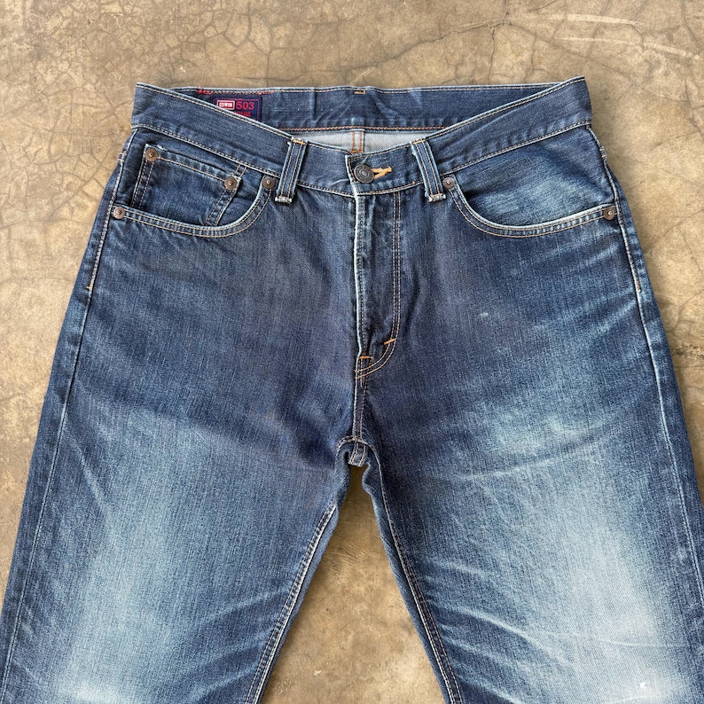 May include: Dark blue denim jeans with a classic five-pocket design. The jeans have a button and zipper closure, with copper-colored rivets and stitching. The denim has a faded appearance, with subtle wear marks.